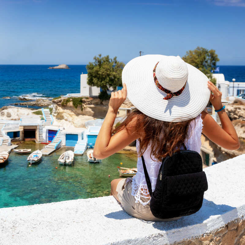 A tourist wearing a big white hat sits on a railing overlooking a charming harbor full of whitewashed houses on an island in Greece, southeast of Europe, Mediterranean region